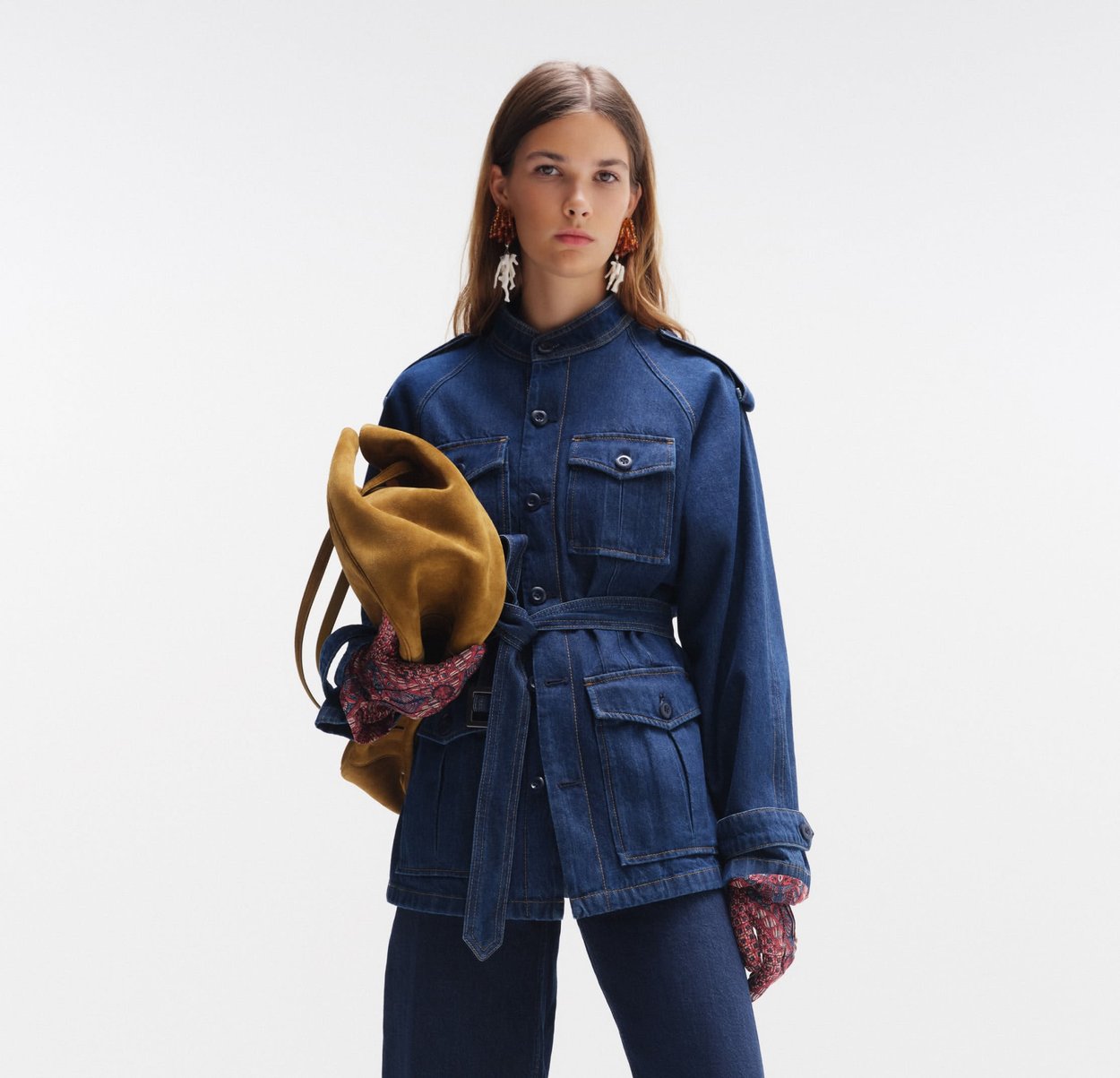 Ung kvinne har på seg en mørk denimjakke med belte og bukser, holder en brun semsket veske, med mønstrede hansker og markante øredobber.