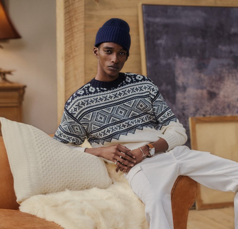 Junger Mann mit einer dunkelblauen Mütze und einem gemusterten Pullover, der auf einem braunen Sofa mit weißen Kissen in einem gemütlichen Raum sitzt.