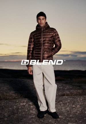 Bruine pufferjack met een rits aan de voorkant, voorzien van een capuchon, gecombineerd met lichtgekleurde broeken. De achtergrond toont een rotsachtig landschap tijdens zonsondergang.