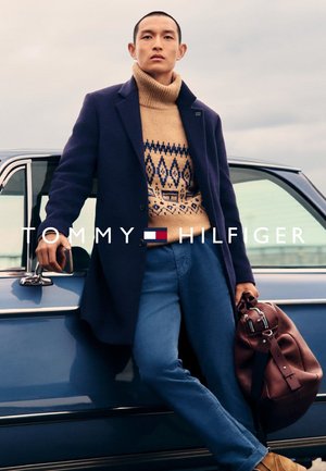 Abrigo de lana azul sobre un suéter de crema con estampado, combinado con pantalones azules. Sosteniendo un bolso de cuero marrón. Con un coche vintage de fondo.