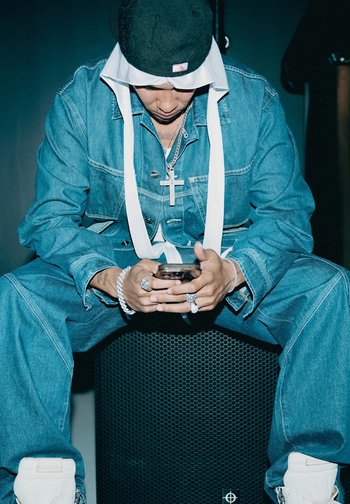 Mann in Jeans-Outfit und Mütze, der auf einem Lautsprecher sitzt, auf sein Handy schaut und eine Kreuzkette, Ringe und Armbänder trägt, im schwachen Licht.
