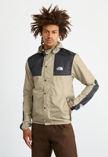 Beige jas met zwarte accenten, drukknopen en capuchon; heeft twee voorzakken, lange mouwen en een logo op de linkerborst en mouwen.