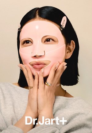 Femme aux cheveux noirs coupés au carré portant un masque facial en tissu rose, tenant son menton avec les deux mains, fond beige, marque Dr.Jart+ en dessous.