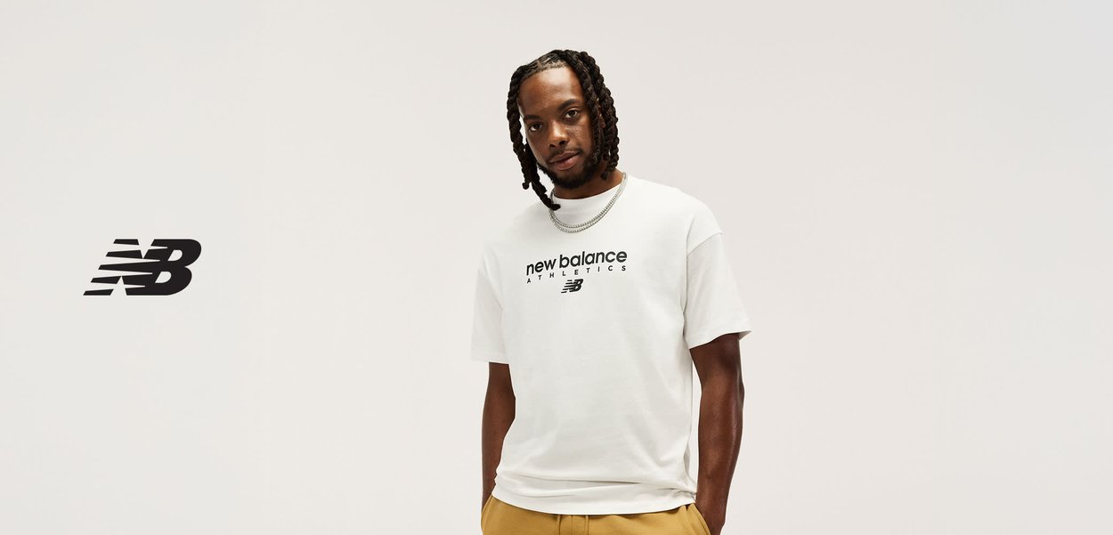 Man met gevlochten haar die een wit New Balance Athletics T-shirt en beige broek draagt, staand tegen een effen lichte achtergrond.