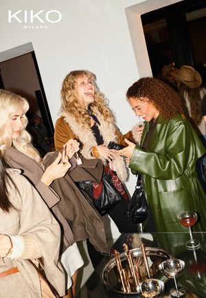 Group of women in various jackets, including a brown fur-accented coat and a green leather jacket, engaging in conversation. A silver tray displays cosmetics.