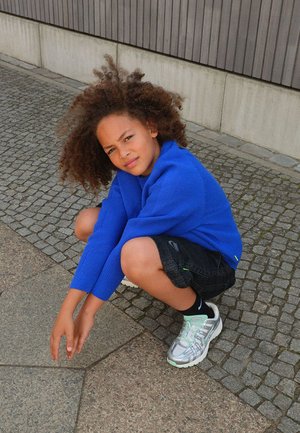 Kind met krullend haar, gekleed in een blauwe trui, zwarte korte broek en zilveren sneakers, hurkend op een betegeld trottoir nabij een betonnen muur.