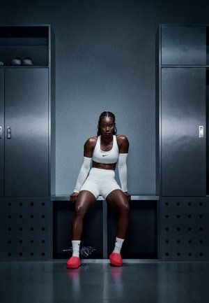 Atleta con reggiseno sportivo e pantaloncini bianchi Nike, che indossa maniche bianche per le braccia e calzature rosso acceso, seduta su una panchina in uno spogliatoio.