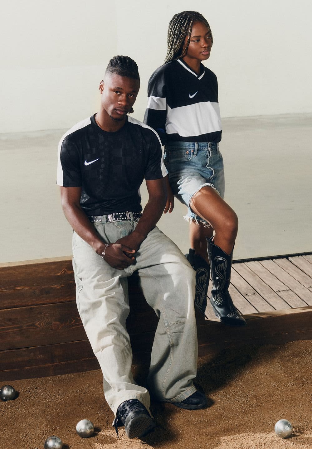 Jersey negro de manga corta con detalles y logo en blanco; pantalones cortos de mezclilla desgastados en lavado ligero; botas de vaquero negras; suelo arenoso, borde de madera.