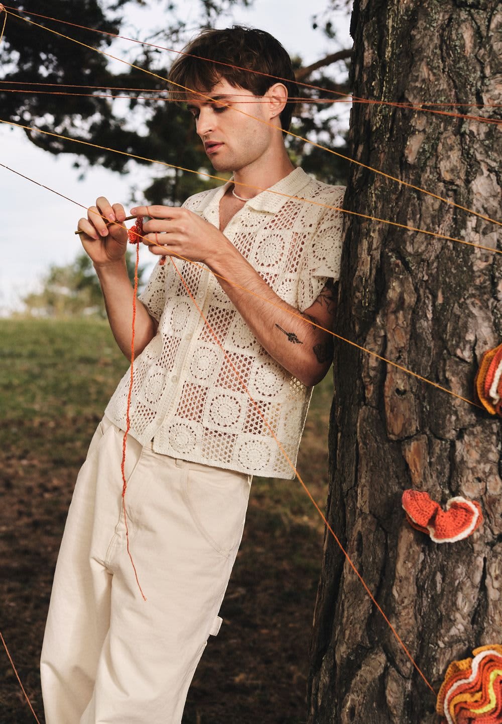 Maglia all'uncinetto di un beige chiaro con motivi a quadrato, abbinata a pantaloni crema. La persona sta lavorando all'uncinetto con filato arancione contro un albero.