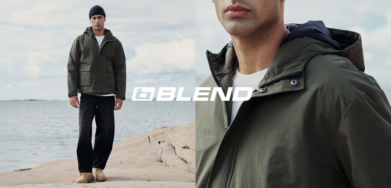 Olivgrüne wasserdichte Jacke mit Kapuze, Fronttaschen und Reißverschluss. Getragen über einem weißen Hemd und schwarzen Hosen, vor einer Küstenlandschaft.