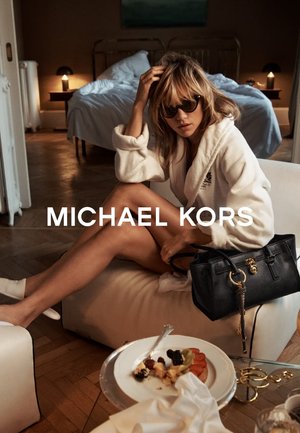 Robe en peluche blanche, sac à main noir avec des accents dorés et lunettes de soleil. Parquet en bois, une assiette avec des fruits et des pâtisseries, et un lit en arrière-plan.