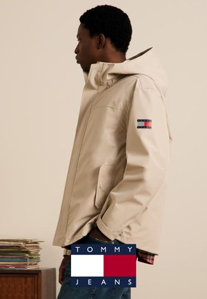 Giacca impermeabile beige con cappuccio, caratterizzata da una toppa con logo sulla manica. Include tasche laterali e una fodera a quadri con bottoni.