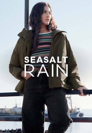 Veste imperméable vert kaki avec capuche, associée à un pull rayé multicolore et un jean noir, sur fond côtier.