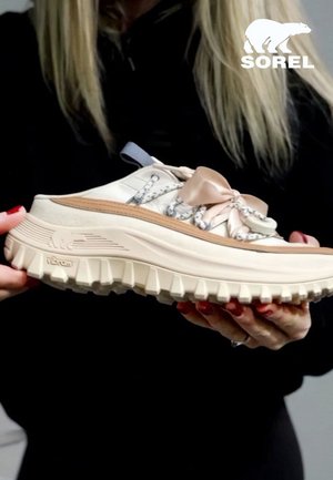 Personne blonde en noir tenant une chaussure SOREL beige et marron clair avec une semelle Vibram épaisse et des lacets ruban devant un fond gris.