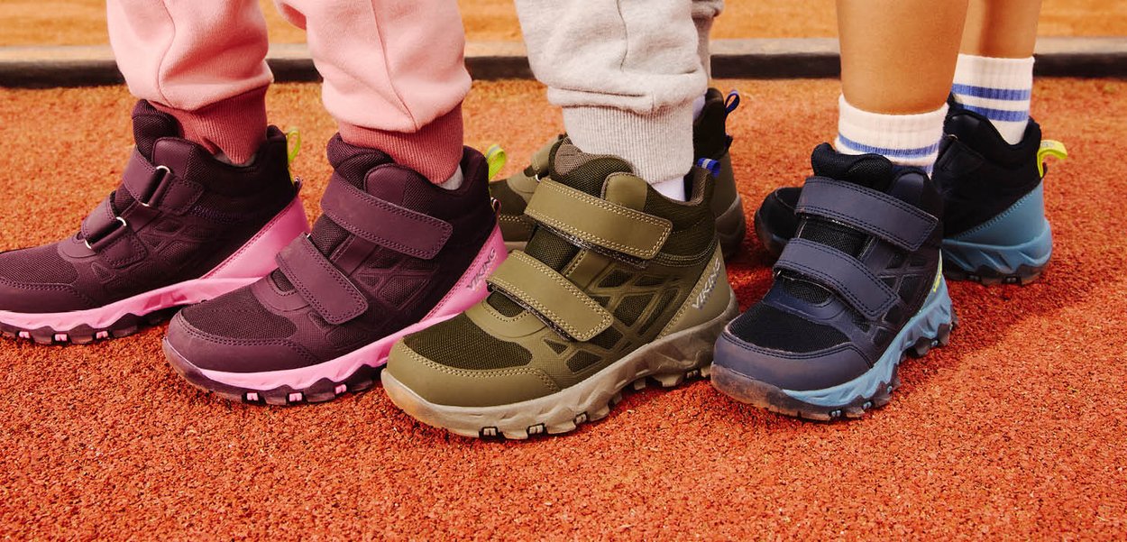 Tre børn iført farverige udendørs sneakers med velcrobånd står tæt sammen på en rød gummibane.