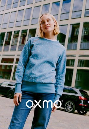 Lichtblauwe sweatshirt met bloemenpatroon, ronde halslijn en schouderknopen. Gecombineerd met een blauwe spijkerbroek. Stedelijke achtergrond.