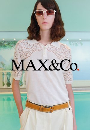 Femme aux cheveux bruns mi-longs portant un polo en dentelle blanche, une ceinture moutarde, un pantalon crème et des lunettes de soleil rectangulaires teintées en rouge, debout près de la piscine.