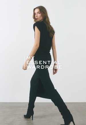 Robe noire ajustée à manches courtes et fente latérale, associée à de hautes bottes noires à talons. Texture lisse sur un fond neutre.