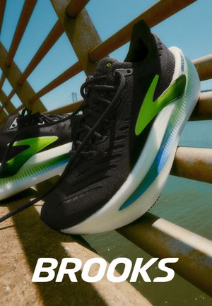 Scarpe da corsa nere con tomaia texturizzata, dettagli verdi e suola con sfumature blu, posizionate davanti a un ponte.