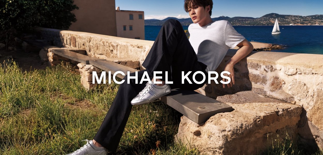 Joven con camisa blanca y pantalones negros relajándose en un banco de piedra junto al mar con un velero de fondo, con el logo de "Michael Kors" superpuesto.