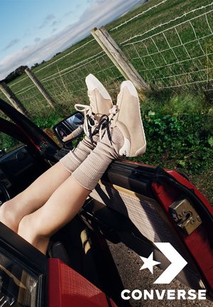 Benen met beige Converse-sneakers en geribbelde sokken steken uit een rode autodeur dichtbij een grasveld met een prikkeldraadhek.