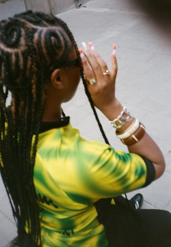 Vlechtfrisuur met gouden accenten, draagt een shirt met korte mouwen in geel en groen, meerdere armbanden om de pols en een zonnebril.