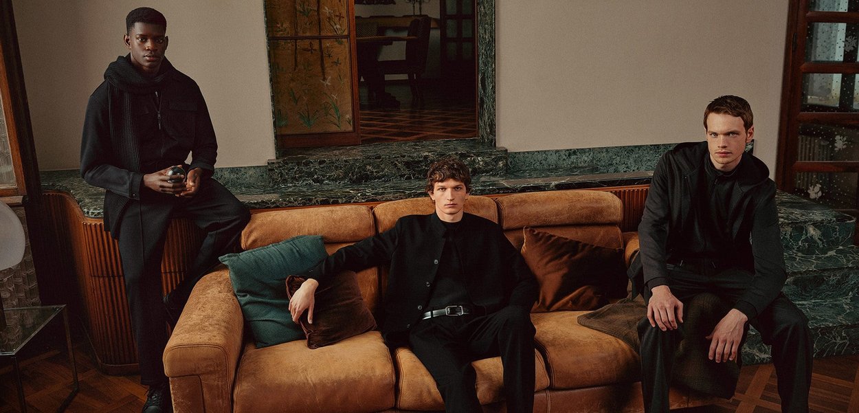 Deux modèles en vêtements sombres sont assis sur un canapé en cuir marron avec des coussins. Le cadre présente un fond en marbre et un sol en bois.