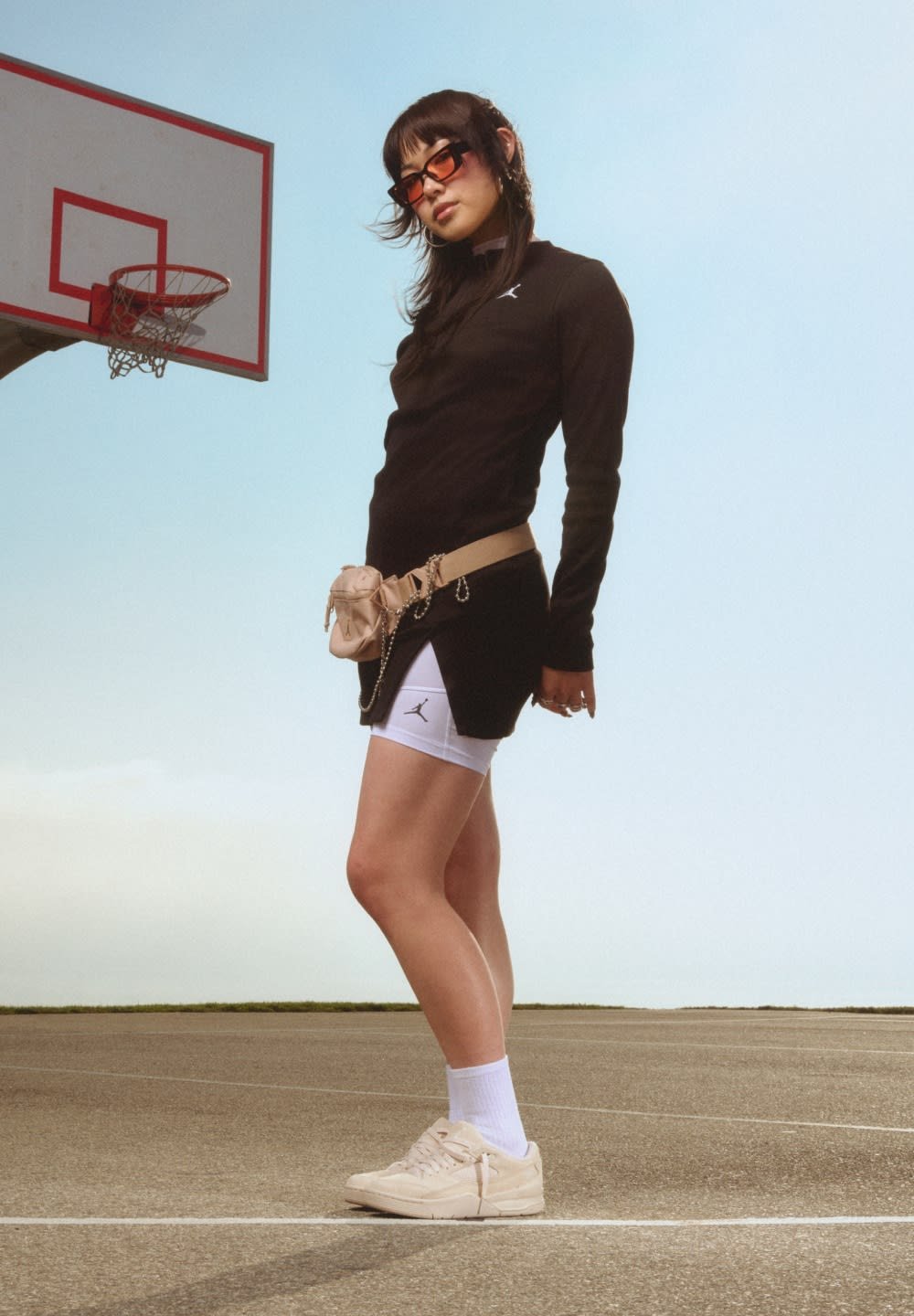 Haut à manches longues noir, shorts blancs avec logo, banane beige clair, et baskets crème. Terrain de basketball en extérieur en arrière-plan.