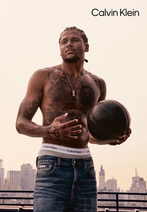Ein Model trägt einen grauen Calvin Klein Bund, blaue Jeans und hält einen schwarzen Basketball. Auf dem nackten Oberkörper sind sichtbare Tattoos zu sehen, vor einer Stadtsilhouette.