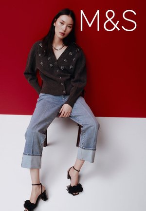 Brązowy dziergany kardigan z ozdobami, jasnoniebieskie spodnie jeansowe do kostek oraz czarne buty na wysokim obcasie z kwiatowymi akcentami na czerwonym tle.