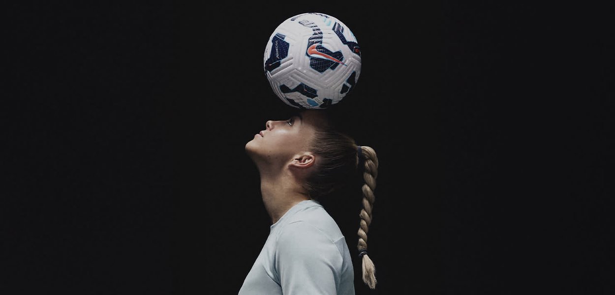 Una pelota de fútbol con una superficie texturizada y acentos azules se equilibra en la cabeza de una persona contra un fondo negro; viste una camiseta de color azul claro.