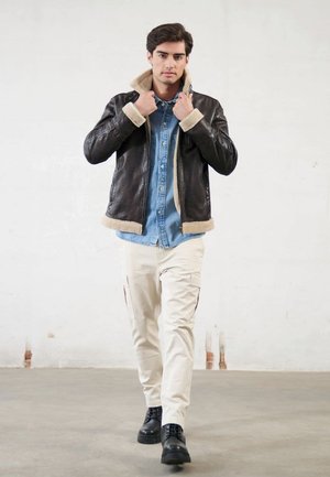 Giacca di pelle marrone con colletto in shearling, camicia di denim blu sotto, pantaloni cargo crema e stivali neri. Modello che cammina in un ambiente interno.