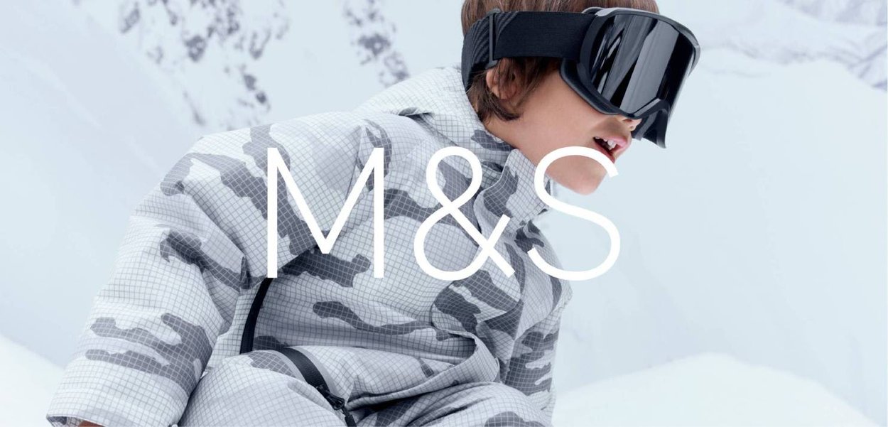 Dziecko ubrane w szary kombinezon zimowy w kamuflażu i czarne gogle narciarskie, w tle zaśnieżone góry.