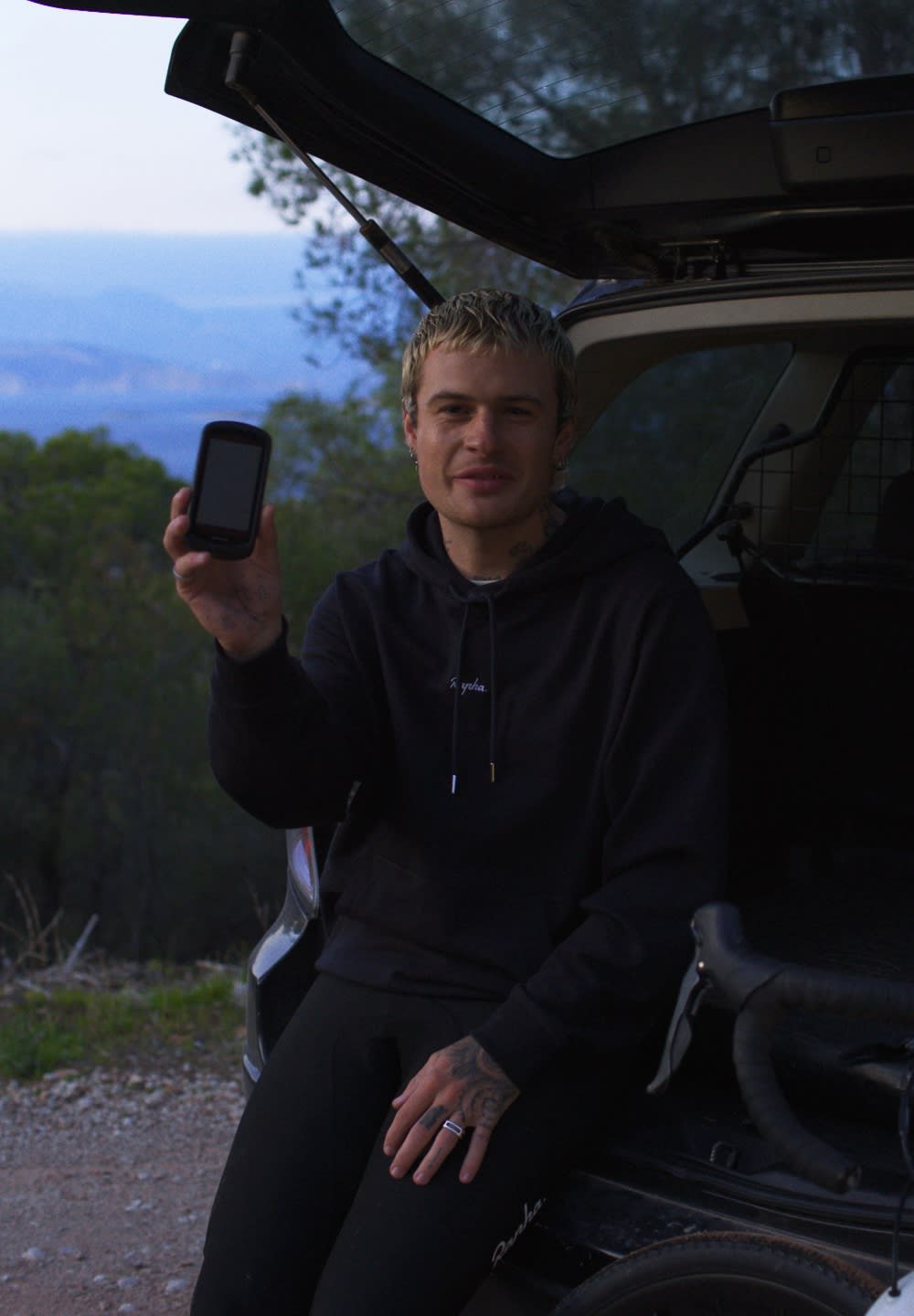 Un téléphone mobile avec une coque noire tenu par une personne dans un sweat à capuche noir, assise dans un véhicule avec un vélo à proximité, un paysage naturel en arrière-plan.
