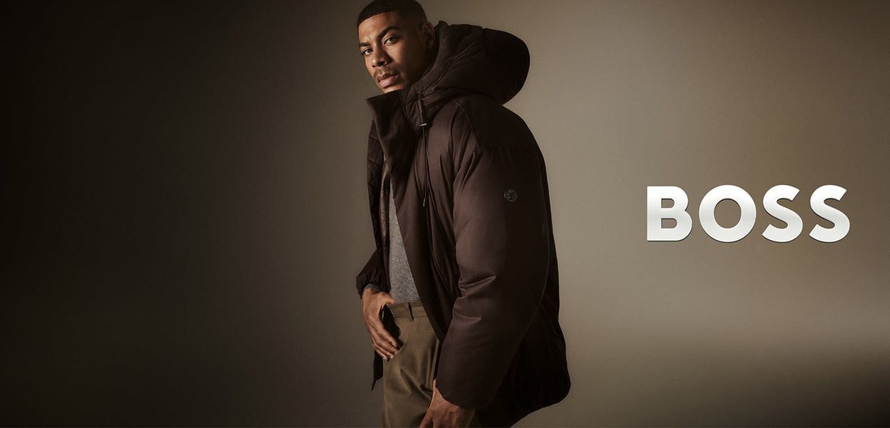 Brown padded jacket with a hood, featuring a zip closure and logo detail. The model wears a gray top and beige pants, set against a neutral background.