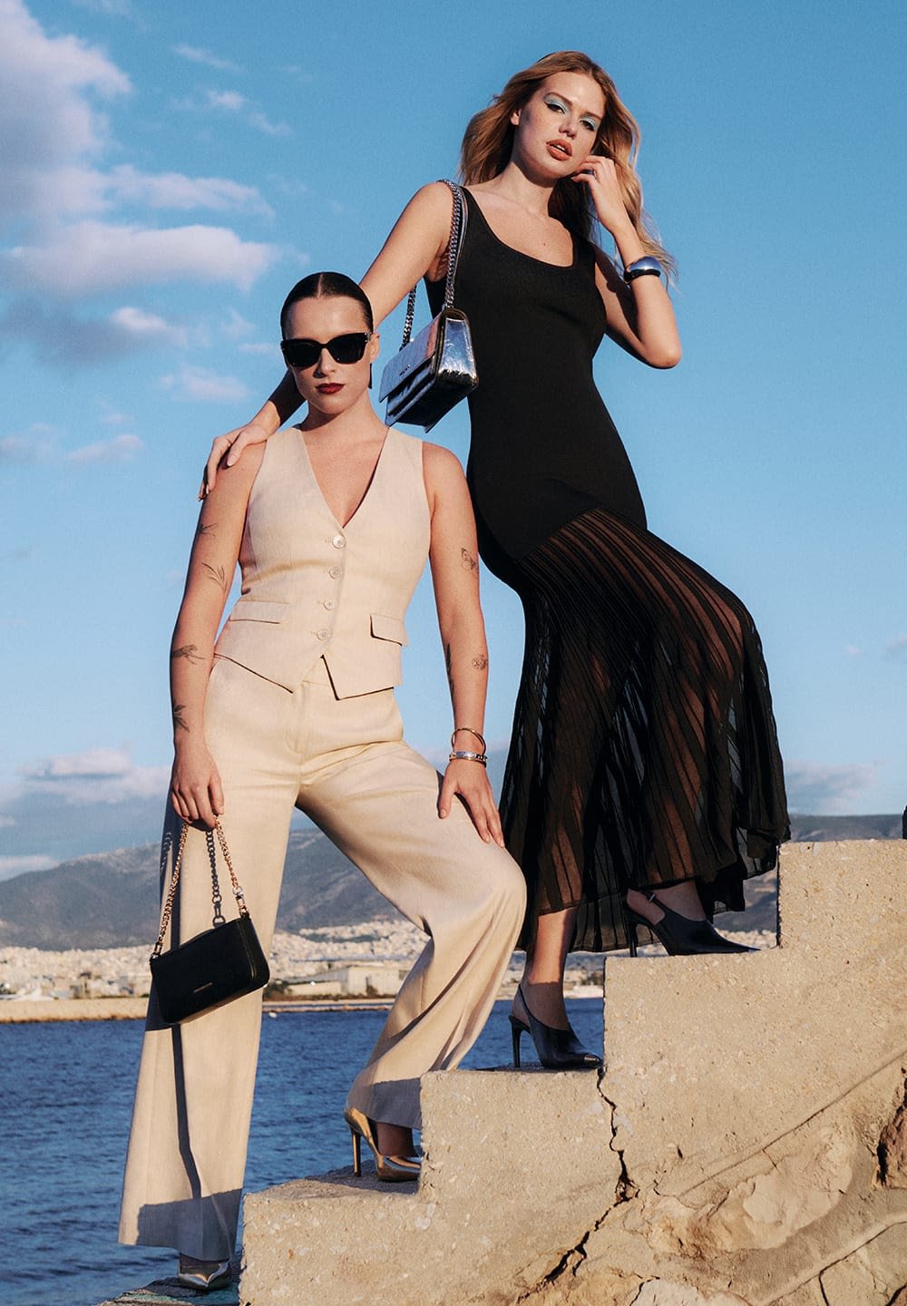 Two women pose on steps beside water. One wears a beige suit and black sunglasses; the other in a black dress with sheer vertical stripes.
