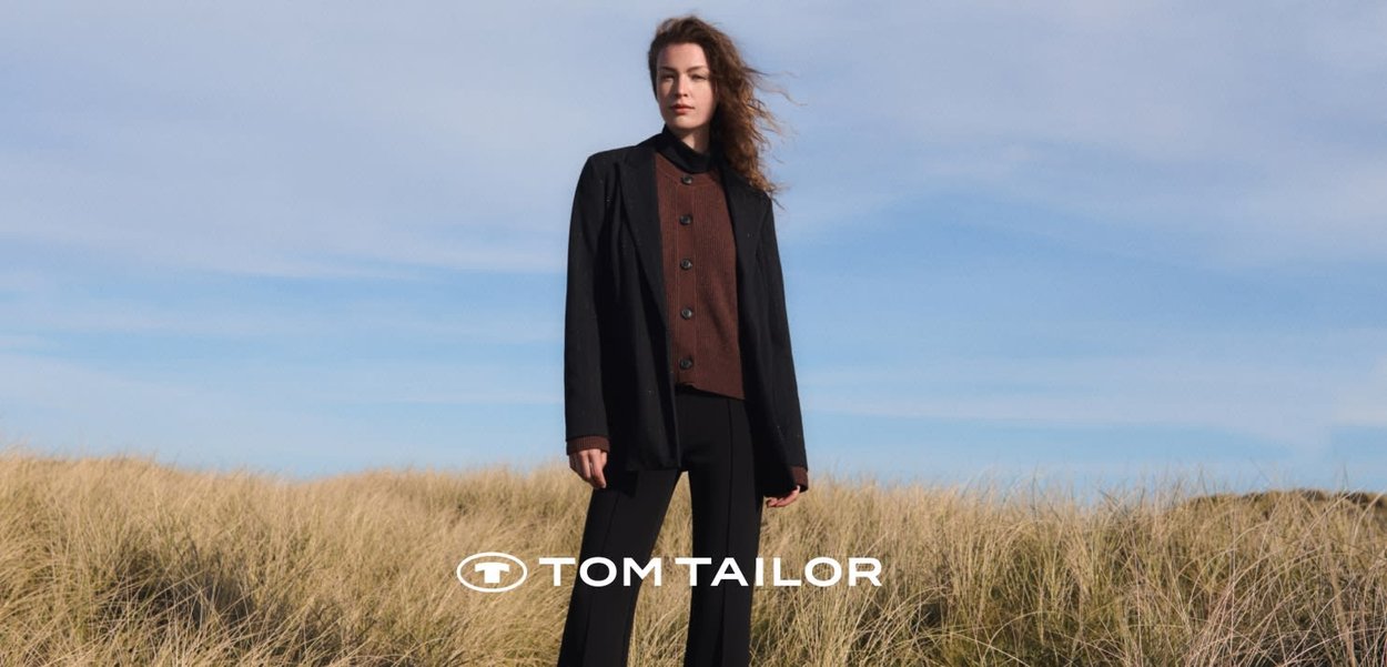 Vrouw staat in een grasveld en draagt een zwart colbert, een bruin vest met knopen en een zwarte broek onder een deels bewolkte blauwe lucht.