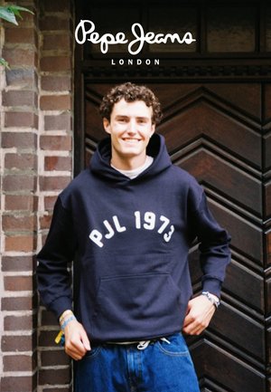 Sweat-shirt à capuche navy avec le texte blanc "PJL 1973", associé à un jean bleu. Tissu texturé avec une coupe décontractée. Mur de briques en arrière-plan.