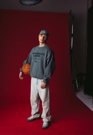 Felpa grigia con il testo "LOS ANGELES LAKERS", jeans azzurri chiari e sneakers grigie, mentre tiene un pallone da basket su uno sfondo rosso.