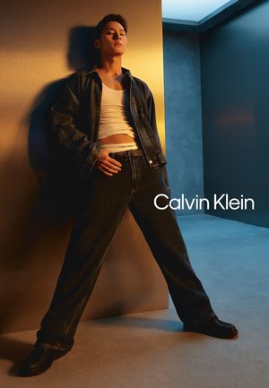 Mann lehnt gegen die Wand, trägt eine Jeansjacke, ein weißes Tanktop und schwarze Jeans, die den Bund seiner Calvin Klein Unterwäsche zeigen.