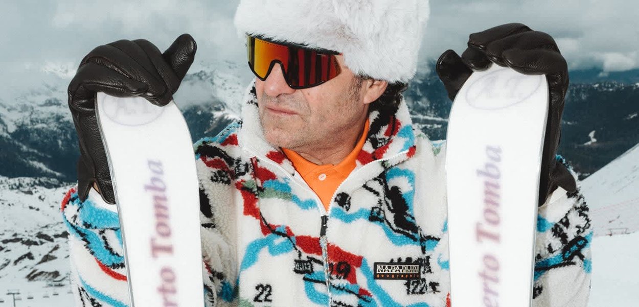 Giacca in pile multicolore con accenti neri, cappello bianco peloso, occhiali da sole specchiati e guanti neri, che tiene sci bianchi con scritte.