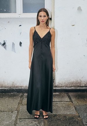 Sort satin maxi-kjole med spaghetti-stropper, prydet med perlebesætninger, en V-udskæring og en flydende silhuet. Båret med sorte hælede sandaler.