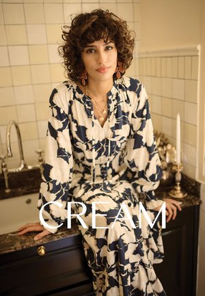 Langärmliges cremefarbenes Kleid mit blauem Blumenmuster, tailliert und mit Rüschen verziert. Vor einem beigen, mit dunklen Akzenten versehenen Fliesenhintergrund.