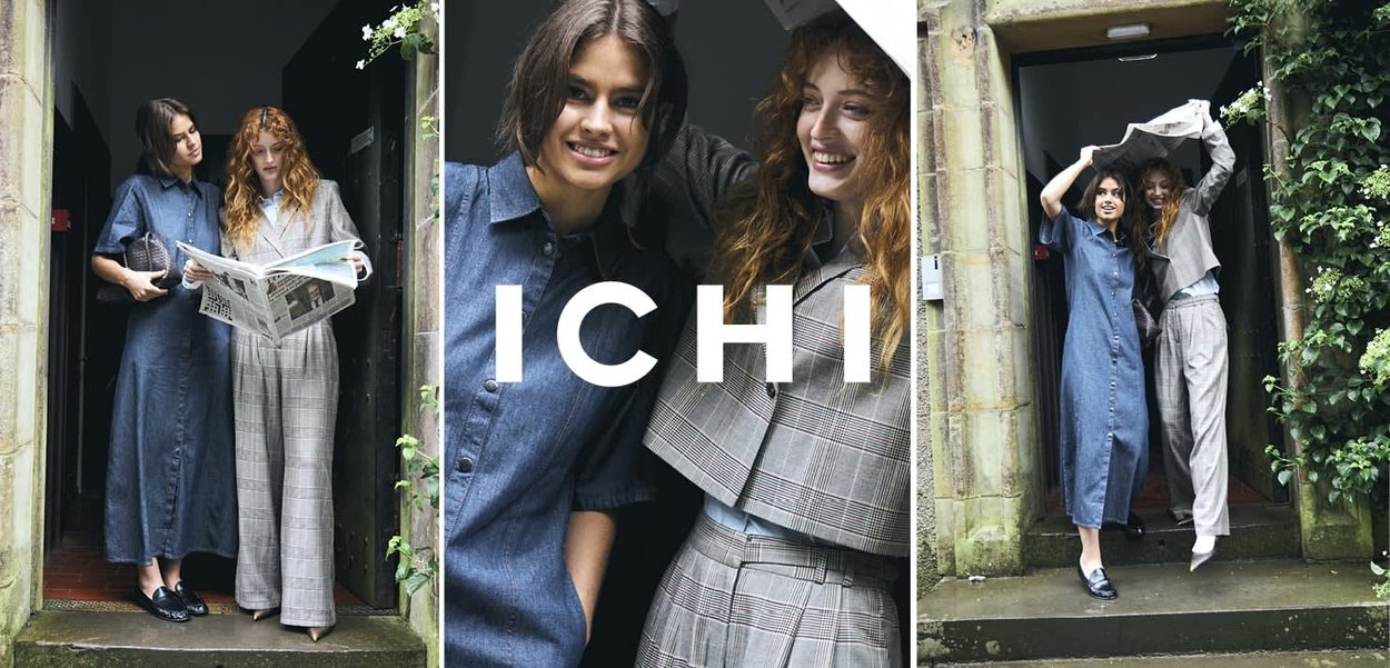 Twee vrouwen staan bij een deur, één leest een krant, beiden dragen stijlvolle casual outfits, glimlachen en hebben interactie buiten.