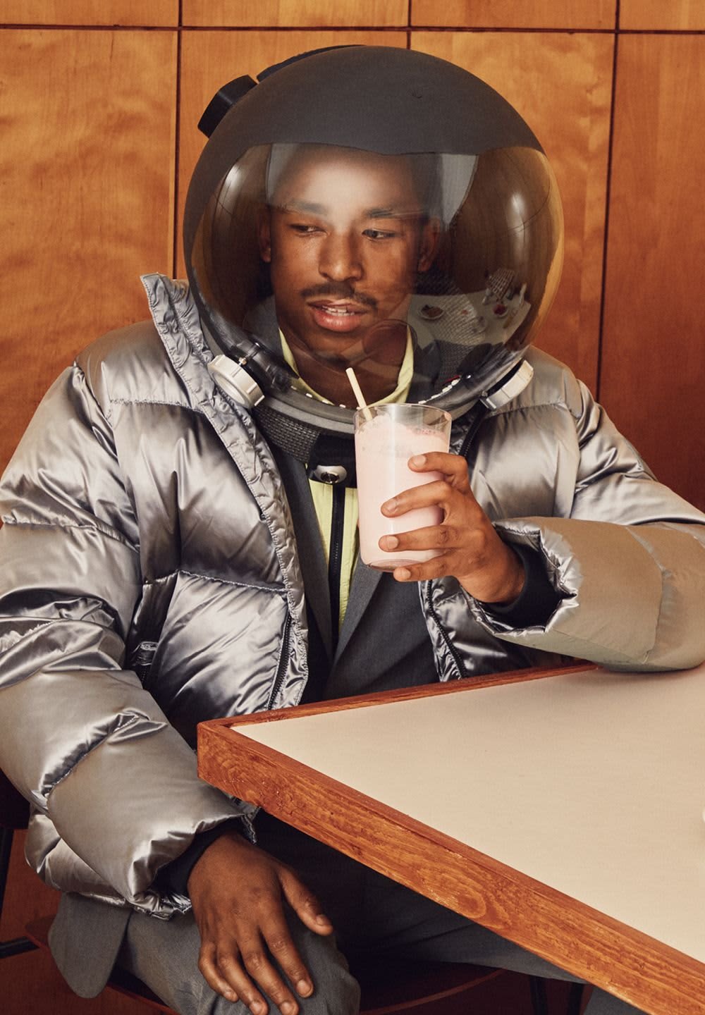 Silver puffer jacket with a round helmet. Holding a glass of pink drink with a straw. Wooden table in the background.