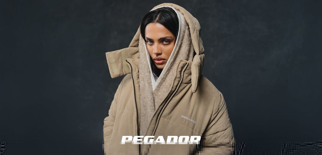 Frau mit dunklen Haaren trägt einen beige übergroßen Kapuzenmantel über einem hellbraunen Hoodie vor einem dunkelgrauen Hintergrund.