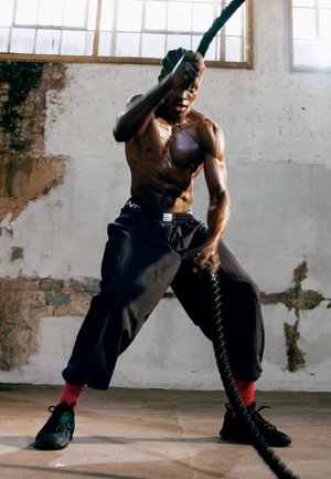 Spierbundel die zware strijdtouwen gebruikt tijdens een training. Hij draagt donkere, loszittende broek en zwarte schoenen met rode accenten.