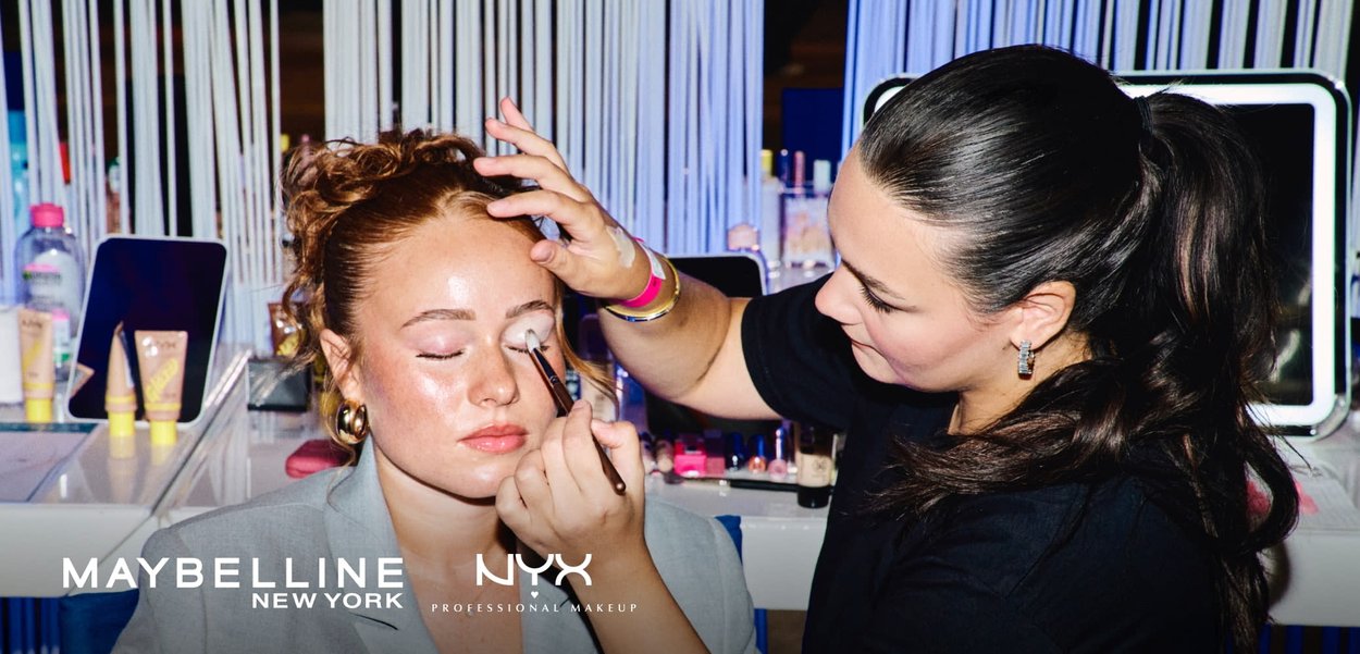 Makeup artist applying eyeshadow on a woman with closed eyes wearing a light gray blazer, with makeup products on a table nearby.