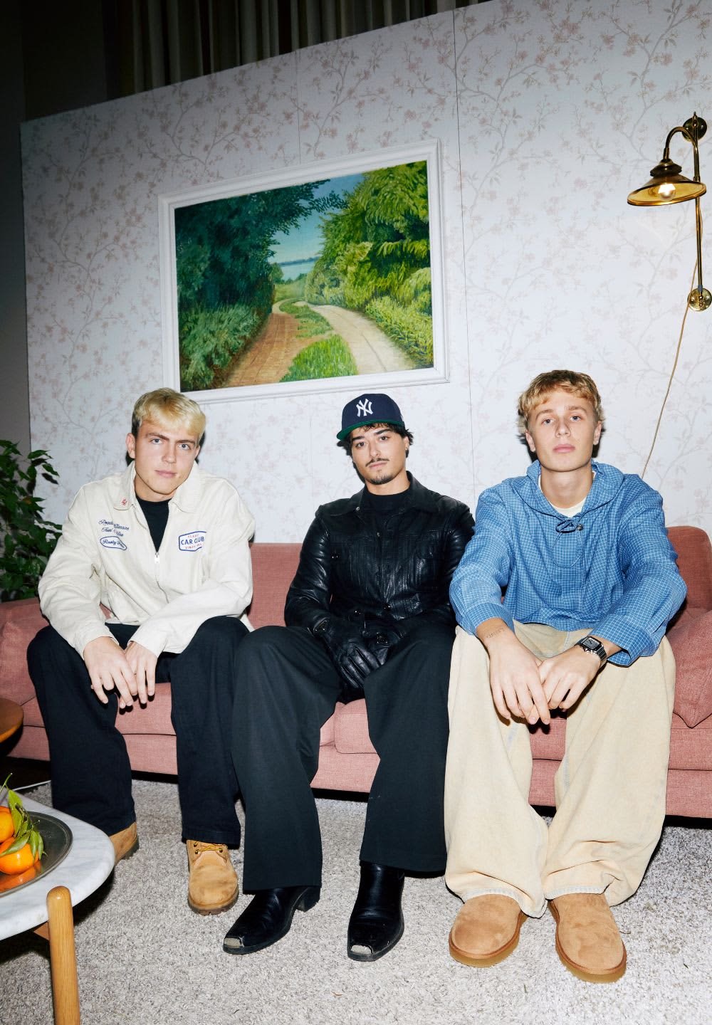 Tre personer sidder på en pink sofa i et rum med blomsterprint på tapetet. De har forskellige stilarter på: en hvid jakke, en sort læderjakke og en blå ternet skjorte.