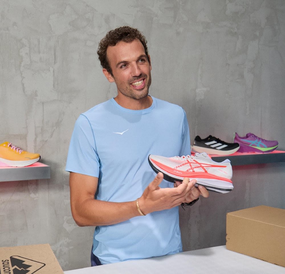 Laufschuh in Grau und Pink, mit einem atmungsaktiven Obermaterial, gepolsterten Sohle und gemusterten Akzenten. Im Hintergrund zusammen mit anderen Schuhen ausgestellt.